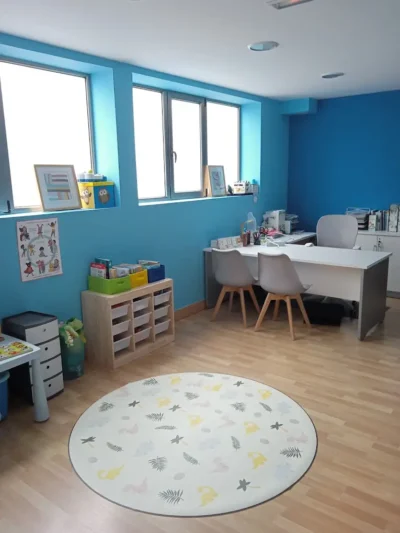 Aula infantil con paredes azules, mesa blanca, sillas y alfombra redonda decorada